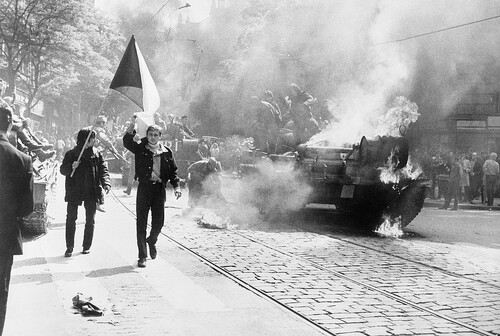 Widok czołgu objętego płomieniami, z którego ewakuuje się załoga. Wokół widoczni przyglądający się ludzie, wśród których na pierwszym planie wyróżniają się dwaj młodzi mężczyźni ze wzniesioną na drzewcu flagą Czechosłowacji.