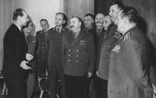 Grupa dowódców w mundurach stojąc w półkolu, wsłuchuje się w słowa mężczyzny w cywilnym ubraniu