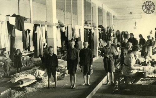 grupka dzieci w ubogich strojach w dużych rozmiarów pomieszczeniu, typu hala., w przestrzeni której widać rozłożone sienniki i rozwieszone części garderoby