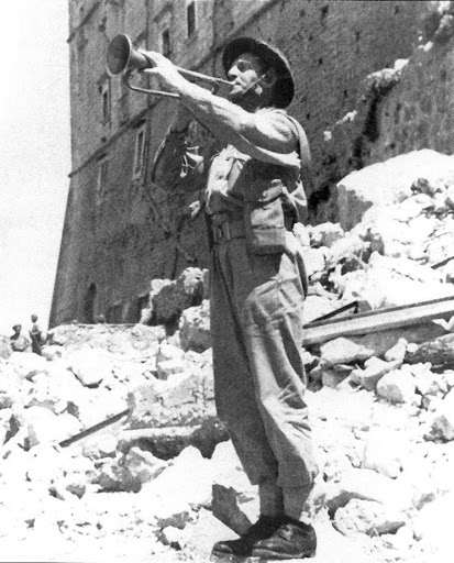Żołnierz w trakcie gry na trąbce w otoczeniu ruin. W tle ściana klasztornego budynku.