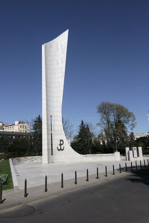 Widok pomnika. Abstrakcyjna konstrukcja przestrzenno-architektoniczna ma postać obelisku w kolorze jasnoszarym. Na nim znak Polski Walczącej.