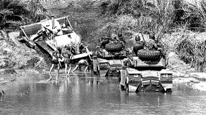 Trzy pojazdy wojskowe wypełnione żołnierzami w trakcie przeprawy przez zbiornik wodny, najprawdopodobniej rzekę