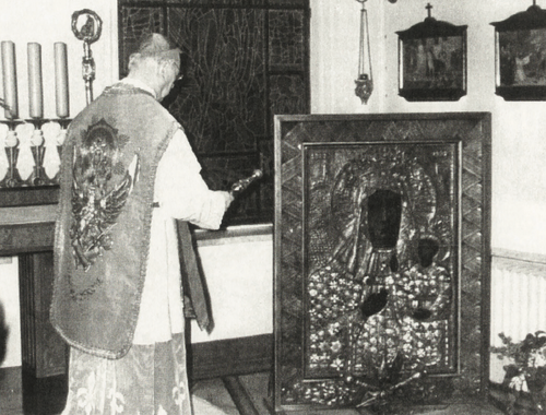 Mężczyzna w stroju liturgicznym, trzymający pastorał w lewej ręce i kropidło w prawej, stoi przed obrazem przedstawiającą Matkę Bożą