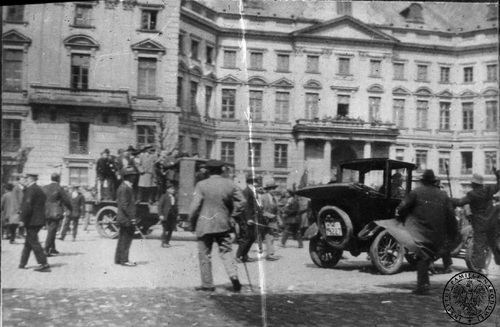 Na archiwalnym zdjęciu grupy ludzi tłoczące się na ulicy w dynamicznych pozycjach. Niektórzy trzymają w ręku przedmioty przypominające laski lub kije. Wśród nich dwa samochody. W tle kamienica.
