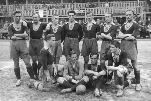 Fotografia drużyny piłkarskiej w strojach sportowych z epoki. W tle zarys trybuny wypełnionej widownią.