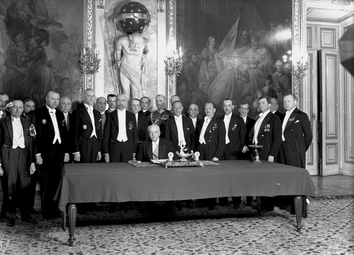 Grupa oficjeli stojąca wokół stołu, przy którym zasiada prezydent. Za nimi widoczne zdobienia wnętrza sali Zamku Królewskiego.
