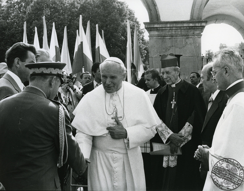 Mężczyzna w białym stroju duchownego katolickiego wita się z mężczyzną w wojskowym mundurze. Wokół stoją osoby w strojach duchownych katolickich. W tle widoczne polskie flagi (biało-czerwone)