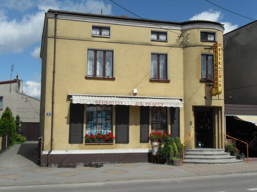 Współczesne zdjęcie budynku. Zabudowa jednopiętrowa ze zmodernizowaną elewacją, na której znajdują się szyldy informujące o działalności sklepu delikatesy w parterowej cześci budynku.