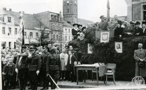 Grupa ludzi zgromadzona wokół trybuny honorowej. Trybuna pokryta igliwiem i udekorowana godłem z Orłem bez korony oraz portretami dwóch osób. Na trybunie oficjele niżej ludność i funkcjonariusze z pocztem sztandarowym.