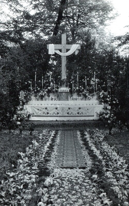 Krzyż ustawiony w miejscu publicznym na tle krzewów, otoczony kościelnymi zdobieniami.