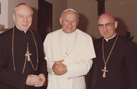 Mężczyzna w białej papieskiej sutannie oraz - po bokach - dwóch mężczyzn w strojach hierarchów katolickich pozują uśmiechnięci do zdjęcia.
