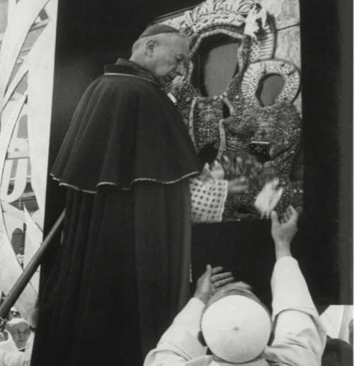 Mężczyzna w stroju hierarchy katolickiego stoi przed obrazem i odbiera od stojącego poniżej zakonnika korny
