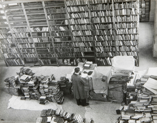 Pomieszczenie biblioteczne. Przed dużym regałem, częściowo wypełnionym książkami, pracownik biblioteki przegląda jedną z książek spośród stosów książek rozstawionych na podłodze.