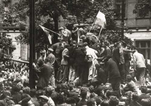 Wśród tłumu zgromadzonego na ulicy, grupa młodych ludzi stoi na platformie czołgu trzymając w ręku polskie flagi.