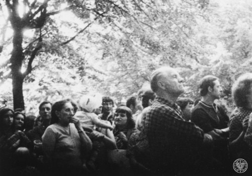 Grupa ludzi, kobiet i mężczyzn, stoi pod drzewami, przypatrując się czemuś lub wsłuchując się w coś z przejęciem. Jedna z kobiet trzyma na rękach małe dziecko.