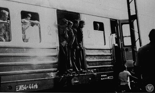 Widać fragment pasażerskiego wagonu kolejowego. W drzwiach wagonu oraz w jego oknach stoją ludzie ubrani w ubrania robocze. Przy wagonie chodzi parę innych osób.