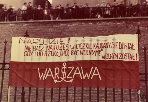 Na białym pasie flagi widoczny napis: &quot;NARODZIE/NIE BACZ NA TO ŻEŚ W CIĘŻKIE KAJDANY SIĘ DOSTAŁ/GDY LUD RZEKŁ &quot;CHCĘ BYĆ WOLNYM!&quot;/WOLNYM ZOSTAŁ&quot;, na tle czerwonym napis: &quot;Warszawa&quot;. W głębi fragment murów klasztoru, na ich szczycie stoją ludzie.