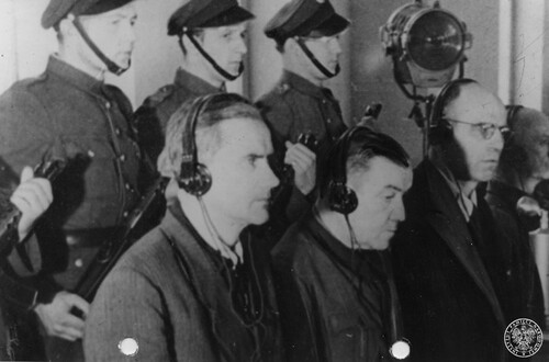 Oskarżeni siedzą ze słuchawkami na uszach; za nimi stoją z bronią maszynową funkcjonariusze