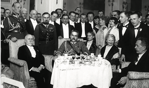Grupa osób w strojach wieczorowych (niektórzy mężczyźni w mundurach) pozują do zdjęcia. Pośrodku, za zastawionym okrągłym stołem, siedzi w fotelu Marszałek Józef Piłsudski