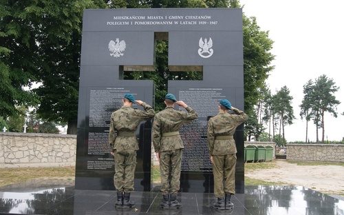Trzech umundurowanych mężczyzn (na zdjęciu stojących tyłem) salutuje prze pomnikiem z czarnego kamienia, w którym pośrodku wyryty został duży krzyż