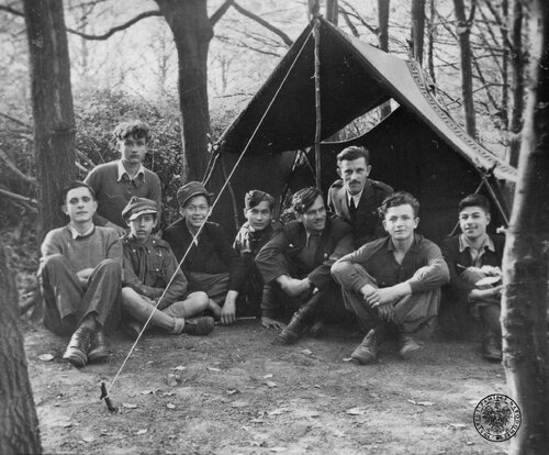 Grupa chłopców, częściowo w mundurach, wraz z jednym dorosłym mężczyzną, siedząca przed wejściem do namiotu