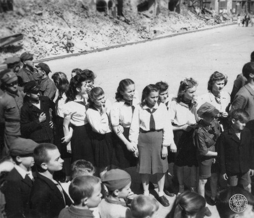 Grupa młodzieży, w centralnej części kilka dziewcząt z zawiązanymi pod szyją chustami (czerwonymi?); w grupie chłopiec w letnim ubraniu harcerskim (czapka, koszula, krótkie spodnie)