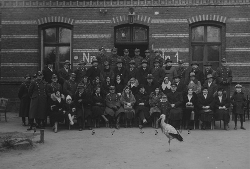 Uczestnicy uroczystości otwarcia świetlicy batalionu morskiego urządzonej przez Polski Biały Krzyż. Wśród siedzących widoczni m.in. ppłk Kurek, dyrektor Wilhelm Urbanicki, ks. dziekan Roszczynalski, kpt. Borysiewicz, porucznikowa Kasztelanowa, Pankowa. Grupa uczestników uroczystości zgromadzona przed wejściem do budynku. W kadrze na pierwszym planie przechadza się bocian.