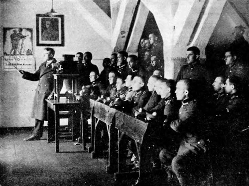 Lekcja historii Polski prowadzona przez Polski Biały Krzyż. Na ścianie obok portretu Józefa Piłsudskiego plakat promujacy PBK. Grupa żołnierzy spoglądających w kierunku obrazu wyświetlanego przez osobę prowadzącą prelekcję.