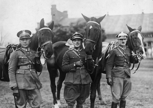 Trójka jeźdźców w mundurach żołnierskich obok koni.