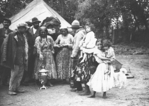 Grupa Cyganów przed namiotem. Jedna z kobiet wskazuje na samowar.