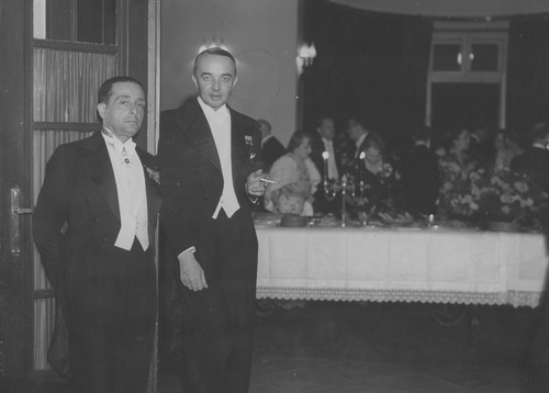 Elegancko ubrani mężczyźni stojący w przejściu do pomieszczenia w którym odbywa się spotkanie w większym gronie.