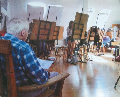 Pracownia malarska, sztalugi, za dwoma z nich widać dwie osoby malujące, z których przynajmniej jedna jest kobietą. Na pierwszym planie, na fotelu, siedzi starszy mężczyzna z siwymi włosami, w okularach i we flanelowej, niebieskiej, w dużą kratę koszuli, trzymając w rękach notatki