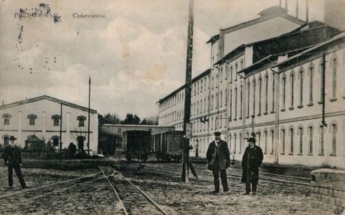 Kompleks budynków, do których prowadzą rozgałęziające się tory kolejowe; na dwóch z nich stoją dwa wagony kolejowe. Na pierwszym planie stoi trzech mężczyzn