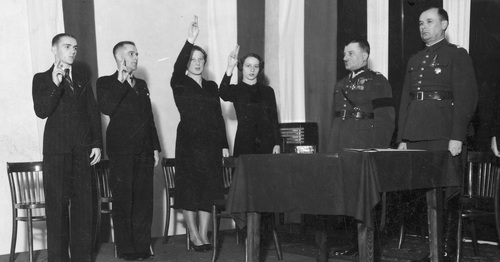 Czwórka sportowców składająca przyrzeczenie, z rękoma uniesionymi w górę, stoją przed dwójką umundurowanych oficerów.