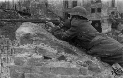 Żołnierz batalionu &amp;quot;Miotła&amp;quot; leżący na gruzach w mundurze i hełmie, w rękach trzyma karabin i celuje.