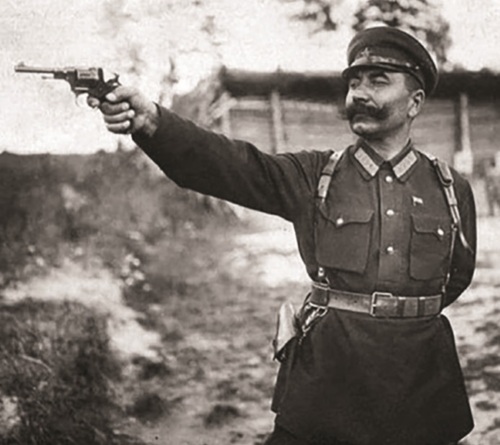 Sowiecki dowódca w mundurze pozuje do zdjęcia z wyciągniętą reką i pistoletem w dłoni.