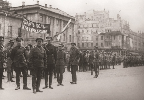 Na ulicy miasta żołnierze w sowieckich mundurach. Dwójka trzyma transparent z hasłem wypisanym cyrylicą.