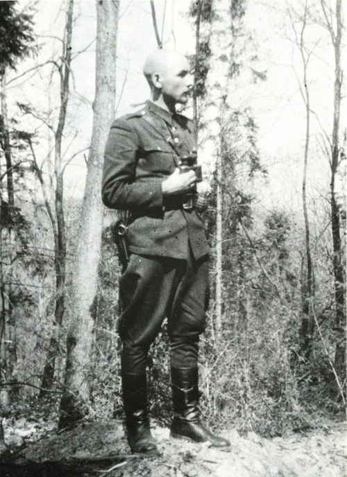 Wysoki mężczyzna, łysy, bez nakrycia głowy, w mundurze wojskowym, stoi wśród rzadkich drzew