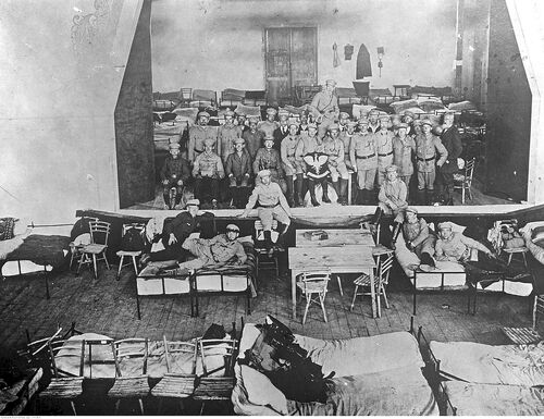 Grupa mężczyzn (najprawdopodobniej w zabudowaniach koszarowych) pozuje do zdjęcia. Z przodu mężczyźni leżą na łóżkach,  kolejni siedzą na krzesłach lub stoją. Zdjęcie wykonane ze znacznego podwyższenia, aby objąć obiektywem jak najwięcej pomieszczenia.