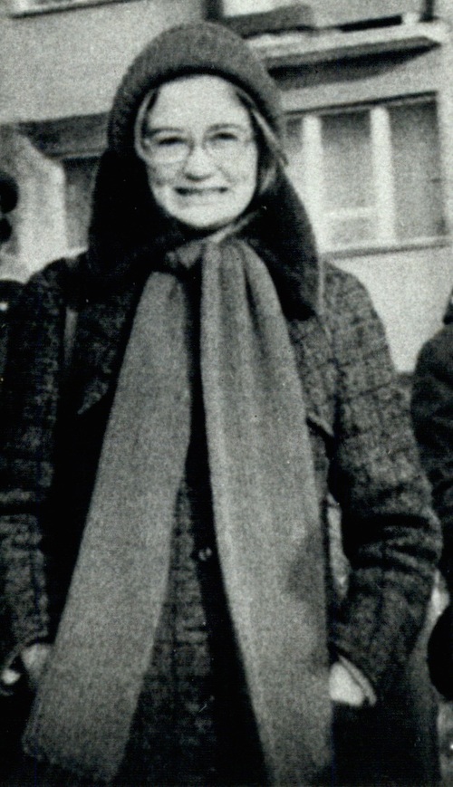 Młoda, szeroko uśmiechnięta kobieta, w okularach z dużymi oprawkami. Kobieta jest ubrana w czapkę, długi szalik i ciepły płaszcz