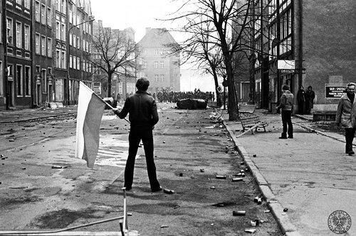 Mężczyzna z flagą Polski stojący samotnie na ulicy Szerokiej. W głębi barykada i kordon milicyjny. Obok idą, stoją, przyglądają się sytuacji nieliczni przechodnie. Po obydwu stronach ulicy budynki