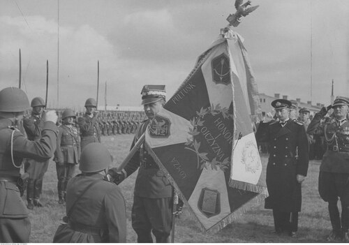 Dowódca w mundurze generalskim przekazuje przyklękającemu żołnierzowi sztandar na którym wypisana jest m.in.  nazwa miasta Łomża.