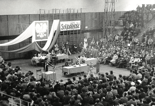 Zdjęcie sali obrad wypełnionej ludźmi. Pośrodku godło PRL i wielka plansza z napisem Solidarność