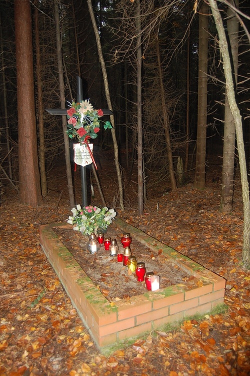 Leśny grób w formie obmurowanej kwatery; nad grobem metalowy krzyż z tabliczką; na grobie znicze i kwiaty