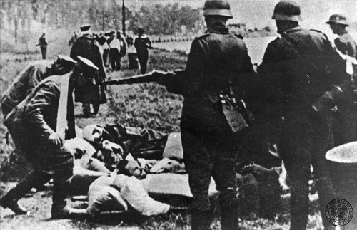 Ciężko ranni członkowie załogi Westerplatte po kapitulacji przed odtransportowaniem do niemieckiego szpitala, 7 września 1939 roku. Na pierwszym planie ranni leżący na noszach na ziemi podnoszeni przez kolegów, obok stoją z bronią pilnujący polskich jeńców żołnierze niemieccy. W tle, pod niemiecką eskortą, maszerują inni jeńcy, w tym żołnierze z zabandażowanymi ranami.