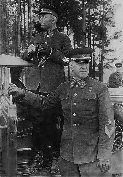 Siemion Timoszenko i Gieorgij Żukow, 1941 rok. Na zdjęciu na pierwszym planie dwóch mężczyzn w średnim wieku w mundurach oficerów Armii Czerwonej. Żukow trzyma lornetkę stojąc na stopniu samochodu osobowego. Timoszenko opiera rękę o okno otwartych drzwi tego samochodu. W głębi drzewa i słabiej widoczny trzeci mężczyzna w mundurze.