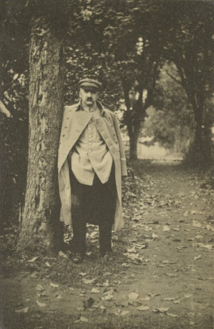 Józef Piłsudski stojący pod drzewem na parkowej alejce. Piłsudski ubrany jest w prostą kurtkę wojskową, zarzucony na ramiona, rozpięty płaszcz wojskowy i czarne spodnie, na głowie ma czapkę-maciejówkę.