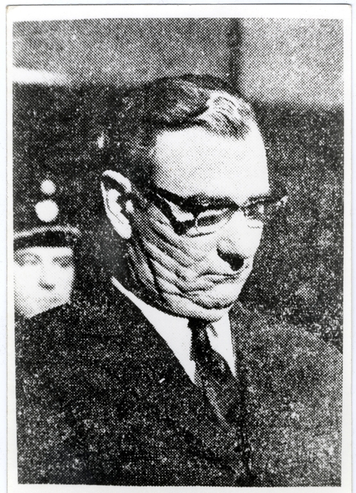 SS-Hupsturmführer Gerulf Mayer podczas rozprawy sądowej w Grazu w 1967 roku. Na zdjęciu jest mężczyzna w średnim wieku, w okularach, gładko ogolony, z dużym podbródkiem, w koszuli, krawacie i marynarce. W tle widać funkcjonariusza policyjnego.