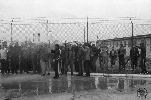 Akcja protestacyjna internowanych w Ośrodku Odosobnienia w Kwidzynie 14 sierpnia 1982 roku. Na zdjęciu widać dużą grupę mężczyzn stojących po dwóch stronach bardzo wysokiego ogrodzenia z metalowej siatki zwieńczonego kilkoma rzędami drutu kolczastego. Mężczyźni zasłaniają się rękami przed strumieniami wody pod ciśnieniem, którą oblewają ich, chcąc spacyfikować protest, służby represyjne Polskiej Rzeczpospolitej Ludowej. Równocześnie większość z mężczyzn ma uniesione do góry dłonie z palcami ułożonymi w znak „V”, symbolizujący wiarę w zwycięstwo nad komunistycznymi prześladowcami. Niektórzy z mężczyzn trzymają w rękach drewniane stołki z wyposażenia Ośrodka. Mężczyźni są ubrani bardzo różnie, niektórzy w dresy sportowe, inni w marynarki lub swetry, paru jest nawet w krótkich spodenkach. W tle podłużny budynek Ośrodka.