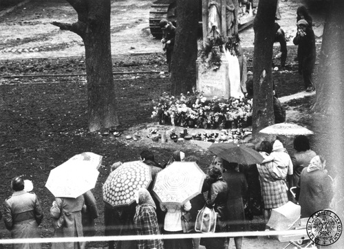 Fotografia wykonana w trakcie obserwacji ludności gromadzącej się pod figurą kamiennego krzyża w Tychach, 1979 r. W tle widoczne gąsienice pojazdu budowlanego. Fot. z zasobu AIPN